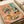 May include: Matisse-inspired still life print displayed in open art book on cream surface with ribbon. Artwork features vibrant coral pink background with wildflower bouquet in glass vase, wine bottle and glass, plates of sliced fruit, bread, cheese, and eggs. Colorful illustration uses bold brushstrokes and simplified forms characteristic of mid-century modern art style.