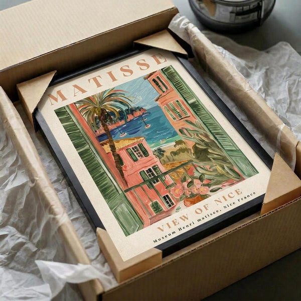 May include: Framed art print featuring a colorful Mediterranean coastal scene with coral-pink buildings, green shutters, palm trees, and turquoise water viewed through an open window. The vintage-style poster shows "MATISSE" and "VIEW OF NICE" text. Black frame sits in cardboard shipping box with protective tissue paper wrapping.