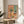 May include: Framed artwork displaying a colorful still life painting with wildflowers in a vase, wine bottle and glass, fruit, and crackers on a pink table against coral and turquoise background, shown leaning on a wooden round side table next to a beige upholstered chair in bright natural light