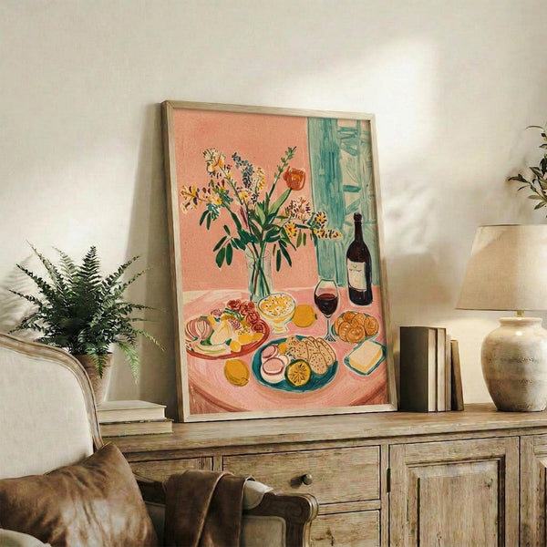 May include: Framed colorful still life artwork displayed on rustic wooden sideboard in bright living room. Print shows pink table with wildflower bouquet in vase, wine bottle and glass, plates of food including cheese and crackers against coral and turquoise background. Surrounded by fern plant, books, ceramic vase, and beige couch with brown leather pillow.