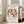 May include: Framed art print featuring Henri Matisse's "The Dance" with five nude figures in terracotta red holding hands in a circle against cream background. Print displays "MATISSE THE DANCE" at top and "BERGGRUEN & CIE" with Paris exhibition details below. Light wooden frame leans against white wall next to beige upholstered sofa in bright, minimalist living room with large windows and natural sunlight streaming across hardwood floors.
