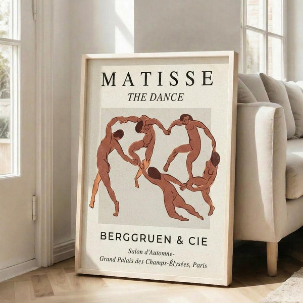 May include: Framed art print featuring Henri Matisse's "The Dance" with five nude figures in terracotta red holding hands in a circle against cream background. Print displays "MATISSE THE DANCE" at top and "BERGGRUEN & CIE" with Paris exhibition details below. Light wooden frame leans against white wall next to beige upholstered sofa in bright, minimalist living room with large windows and natural sunlight streaming across hardwood floors.