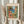 May include: Person holding framed art print in light wooden frame showing colorful Mediterranean coastal scene with coral-pink buildings, green shutters, palm tree, blue water with boats, and tropical plants. Dried pampas grass visible on left side against neutral curtained background.