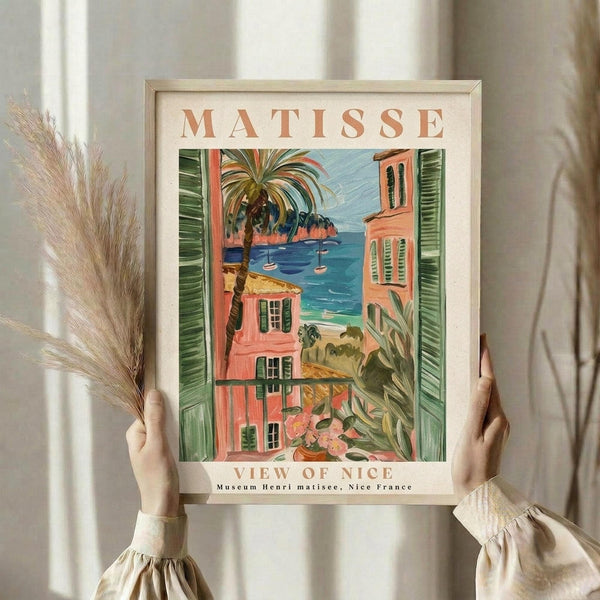 May include: Person holding framed art print in light wooden frame showing colorful Mediterranean coastal scene with coral-pink buildings, green shutters, palm tree, blue water with boats, and tropical plants. Dried pampas grass visible on left side against neutral curtained background.