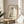 May include: Framed minimalist artwork featuring a sleeping gray cat drawn in Japanese ink wash style with black calligraphy characters above, displayed on a round wooden side table next to a beige upholstered chair with white throw pillow, green plant leaves visible in corner, natural sunlight creating soft shadows on neutral walls