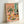 May include: Framed art print leaning against white wall showing colorful still life scene with wildflower bouquet in glass vase, wine bottle and glass, plates of food including sliced eggs and bread, all arranged on pink tablecloth. Features coral pink background, turquoise curtains, and vibrant colors in an illustrative style reminiscent of mid-century modern art.