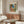 May include: Matisse poster print displayed above wooden sideboard in modern living room. The colorful artwork shows Mediterranean coastal scene with coral buildings, green shutters, palm trees, and blue ocean view. White curved chair and glass coffee table complete the mid-century modern interior styling.
