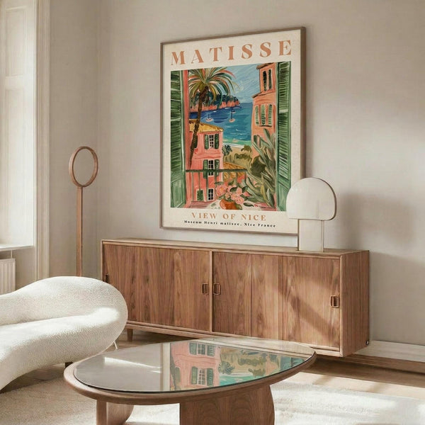 May include: Matisse poster print displayed above wooden sideboard in modern living room. The colorful artwork shows Mediterranean coastal scene with coral buildings, green shutters, palm trees, and blue ocean view. White curved chair and glass coffee table complete the mid-century modern interior styling.