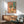 May include: Colorful still life poster with coral pink background showing wildflowers in glass vase, wine bottle, glass of red wine, plates of food and crackers, displayed in thin wooden frame above mid-century modern walnut credenza with white ceramic lamp, next to sage green upholstered chair with wooden arms in bright white room with window shadows.