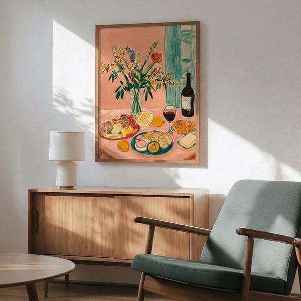 May include: Colorful still life poster with coral pink background showing wildflowers in glass vase, wine bottle, glass of red wine, plates of food and crackers, displayed in thin wooden frame above mid-century modern walnut credenza with white ceramic lamp, next to sage green upholstered chair with wooden arms in bright white room with window shadows.
