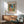 May include: Framed Matisse poster showing colorful Mediterranean coastal scene with coral buildings, green shutters, palm trees, and blue water, displayed above wooden sideboard with lamp in modern living room setting with sage green mid-century chair and natural lighting creating wall shadows.