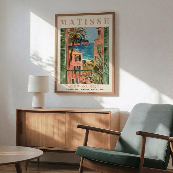 May include: Framed Matisse poster showing colorful Mediterranean coastal scene with coral buildings, green shutters, palm trees, and blue water, displayed above wooden sideboard with lamp in modern living room setting with sage green mid-century chair and natural lighting creating wall shadows.