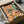 May include: Colorful still life artwork in black frame being unboxed from cardboard packaging with tissue paper. The illustration features a coral pink background with wildflower bouquet in glass vase, wine bottle and glass, assorted fruits, cheese, and crackers arranged on a table. Green curtains visible in background. Matisse-inspired flat illustration style with bold colors and simplified forms.