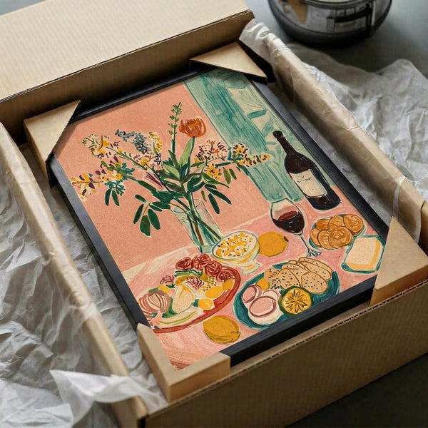 May include: Colorful still life artwork in black frame being unboxed from cardboard packaging with tissue paper. The illustration features a coral pink background with wildflower bouquet in glass vase, wine bottle and glass, assorted fruits, cheese, and crackers arranged on a table. Green curtains visible in background. Matisse-inspired flat illustration style with bold colors and simplified forms.