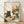 May include: Framed art print showing a gray tabby cat sitting at a table with checkered tablecloth, yellow coffee cup, wooden mug, open book, and large floral bouquet with sunflowers and pink blooms in green vase. Natural wood frame leaning against white textured wall on light wooden floor with diagonal window shadows.