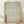 May include: Framed poster with traditional Japanese wave pattern in sage green lines on cream background, featuring repeating ocean waves and stylized water motifs. Dark red Japanese characters appear in bottom left corner. Natural wood frame leans against white wall on light wooden floor with soft natural lighting casting shadows.