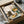 May include: Framed art print in black frame being unwrapped from cardboard shipping box with protective paper. Illustration shows dark gray tabby cat sitting on wooden chair at checkered tablecloth table with yellow coffee cup, sunflowers and colorful wildflowers in green vase. Warm cream background with soft window light shadows. Cottage core style artwork with muted yellows, greens, and earth tones.