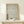 May include: Framed artwork featuring intricate blue-green wave patterns in traditional Japanese style against cream background, with vertical red Japanese characters on left side. Natural wood frame sits on light wooden console table beside small white wrapped gift with evergreen sprig. Soft lighting creates gentle shadows on neutral wall.