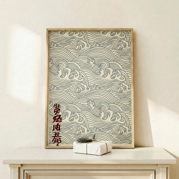 May include: Framed artwork featuring intricate blue-green wave patterns in traditional Japanese style against cream background, with vertical red Japanese characters on left side. Natural wood frame sits on light wooden console table beside small white wrapped gift with evergreen sprig. Soft lighting creates gentle shadows on neutral wall.