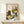 May include: Framed poster print on light wooden shelf showing black cat with golden eyes sitting at checkered tablecloth table with yellow coffee cup, colorful flower bouquet in green vase featuring sunflowers and pink blooms, open book, all in cozy illustrated art style. Small wrapped gift with sprig sits on shelf beside frame against cream wall with natural window lighting.