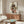 May include: Framed reproduction of early 20th century expressionist artwork showing two figures in dark formal wear against cream background, displayed above walnut wood credenza in modern living room with white curved chair, brass floor lamp, oval glass coffee table, and neutral walls with natural lighting