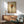 May include: Framed poster print featuring pasta wrapped around a fork against a beige background, displayed above a wooden credenza in a modern living room with mid-century furniture, white table lamp, and sage green upholstered chair. Natural sunlight creates geometric shadows on white walls.