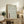 May include: Framed artwork featuring intricate wave patterns in sage green lines on cream background with red Japanese characters, displayed on rustic wooden sideboard alongside fern plant, ceramic vase, and books in neutral-toned living room setting