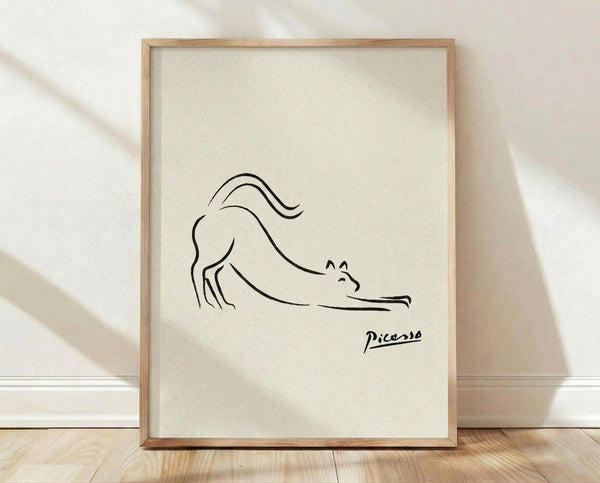 May include: Minimalist line drawing of a stretching cat in black ink on cream textured paper, displayed in light wooden frame against white textured wall with soft shadows. Simple continuous line art shows feline in downward stretch pose with curved tail, signed "Picasso" in bottom right corner.