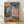 May include: Framed reproduction of Picasso's 1905 painting showing two seated acrobats - a young boy in diamond-patterned harlequin costume and white ruffled collar next to an older figure in pink garment with white collar. The artwork features muted blues, pinks, and earth tones characteristic of Picasso's Rose Period. Natural wood frame displays against white wall with sunlight casting soft shadows on light wooden floor.