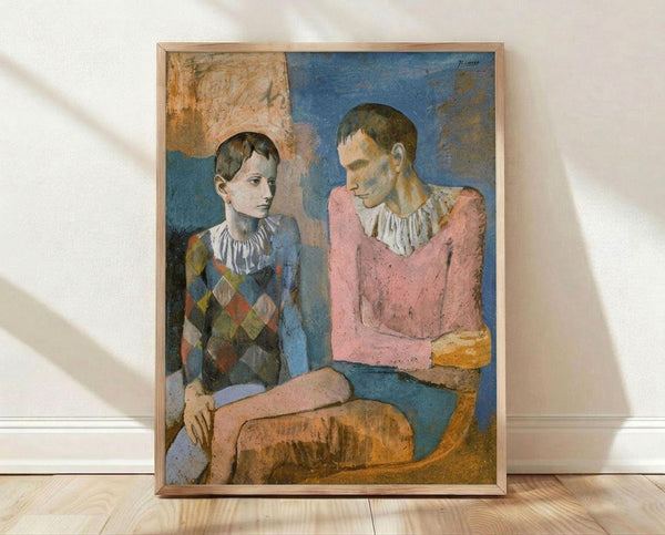 May include: Framed reproduction of Picasso's 1905 painting showing two seated acrobats - a young boy in diamond-patterned harlequin costume and white ruffled collar next to an older figure in pink garment with white collar. The artwork features muted blues, pinks, and earth tones characteristic of Picasso's Rose Period. Natural wood frame displays against white wall with sunlight casting soft shadows on light wooden floor.