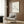 May include: Minimalist line drawing of a stretching cat with Picasso signature on cream-colored poster in light wood frame, mounted on rich walnut wood paneling. Beige upholstered armchair with geometric patterned pillow sits below artwork, alongside brass floor lamp and dark side table with open book. Neutral curtains frame window in modern living room setting.