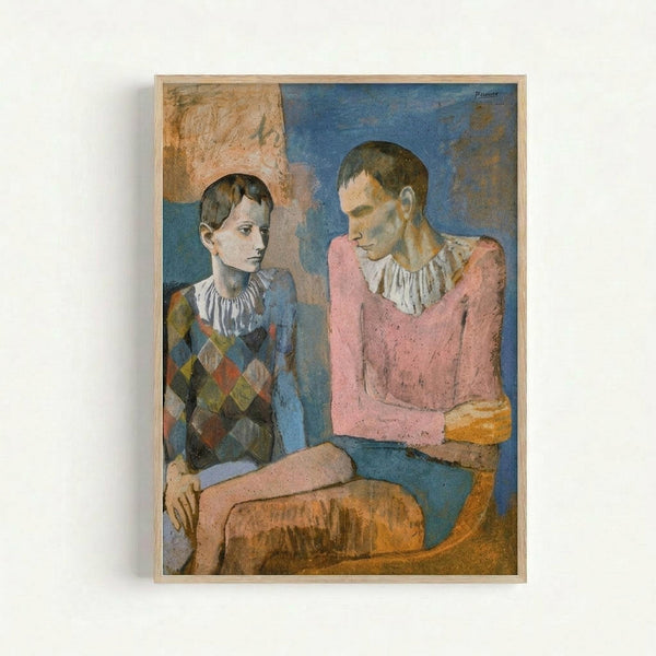 A framed reproduction of Picasso's painting showing two young acrobats sitting together. The boy on the left wears a diamond-patterned harlequin costume in muted colors, while the figure on the right wears a pink garment with white collar details. The painting features Picasso's early period style with soft blue and earth-tone backgrounds. Both figures have contemplative expressions and are rendered in the artist's characteristic simplified forms. The artwork is displayed in a light wooden frame