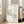May include: Minimalist line drawing of a stretching cat in black ink on cream background, displayed in light wooden frame leaning against white wall next to beige upholstered chair. Sunlight streams through tall windows onto hardwood floors in bright, airy living room setting.