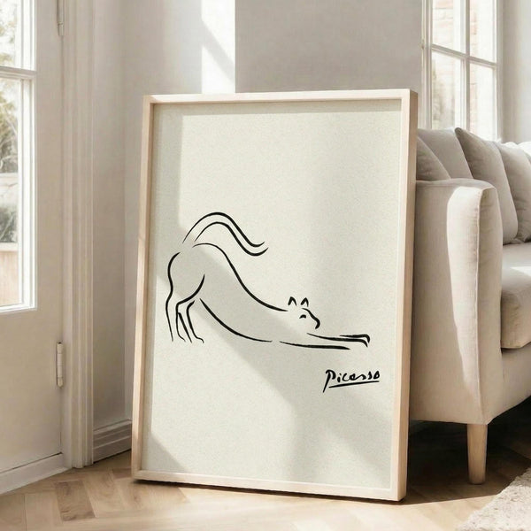 May include: Minimalist line drawing of a stretching cat in black ink on cream background, displayed in light wooden frame leaning against white wall next to beige upholstered chair. Sunlight streams through tall windows onto hardwood floors in bright, airy living room setting.