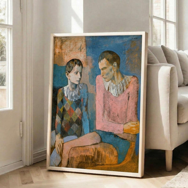 May include: Framed reproduction of Picasso's 1905 painting showing two young acrobats in harlequin costumes sitting together. The left figure wears a diamond-patterned outfit in blues and earth tones, while the right wears a pink top. The painting features muted blues, pinks, and golden yellows with Picasso's characteristic early style. Large framed print leans against a light wooden chair in a bright, minimalist room with white walls and windows.
