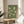 May include: Framed poster with sage green background featuring white cranes in flight, displayed on round wooden side table next to beige upholstered chair. Natural lighting creates soft shadows across cream walls, with plant leaves visible in corner of minimalist living room setting.
