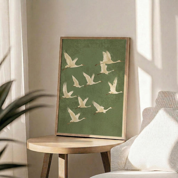 May include: Framed poster with sage green background featuring white cranes in flight, displayed on round wooden side table next to beige upholstered chair. Natural lighting creates soft shadows across cream walls, with plant leaves visible in corner of minimalist living room setting.