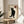 May include: Framed poster print featuring a black cat with yellow eyes sitting upright on cream-colored paper background, with vertical Japanese kanji characters on the left side and a red heart containing a kanji character in the upper right corner. The wooden-framed artwork sits on a light wood round side table next to a beige upholstered chair, with a potted plant visible in soft natural lighting.