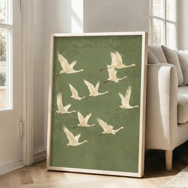 May include: Framed art print featuring white cranes in flight against a textured sage green background, displayed in a bright living room setting next to a beige upholstered chair. The artwork shows multiple birds with outstretched wings in various flying poses, housed in a natural wood frame and leaning against white walls with large windows.
