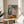 May include: Framed reproduction of Picasso's 1905 painting showing two seated acrobats in harlequin costumes with white ruffled collars - one in diamond-patterned blue outfit, one in pink. The poster sits on a light wood round side table next to a beige upholstered chair with white pillow. Natural sunlight streams across white walls creating geometric shadows. Green plant leaves visible in corner.