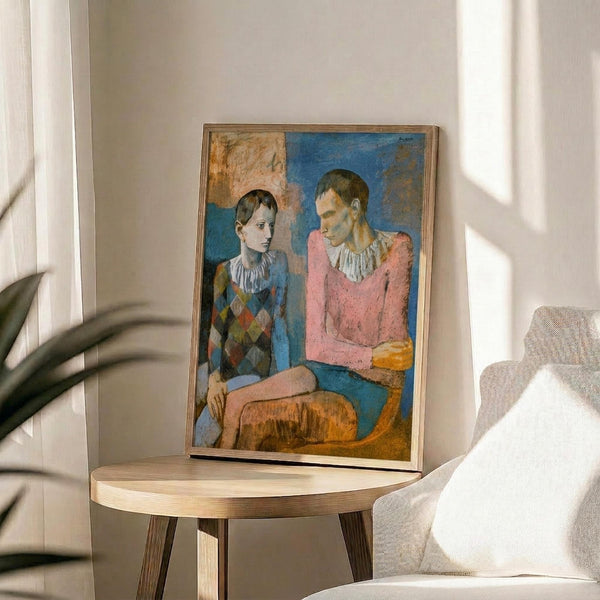 May include: Framed reproduction of Picasso's 1905 painting showing two seated acrobats in harlequin costumes with white ruffled collars - one in diamond-patterned blue outfit, one in pink. The poster sits on a light wood round side table next to a beige upholstered chair with white pillow. Natural sunlight streams across white walls creating geometric shadows. Green plant leaves visible in corner.