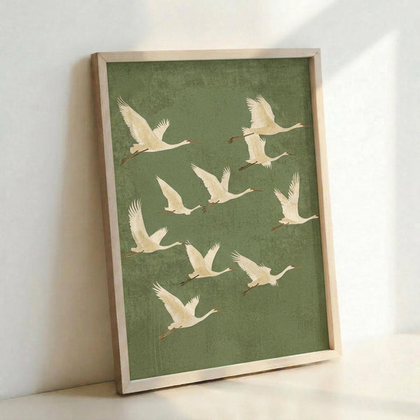May include: Framed wall art print featuring nine white cranes in flight against a muted sage green textured background, displayed in a light wood frame leaning against a white wall. The birds are arranged in a flowing diagonal pattern with wings spread and long necks extended, showing delicate feather details and orange-red legs.