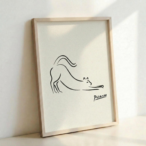 May include: Minimalist line drawing of a stretching cat in black ink on cream paper, displayed in a light wood frame leaning against a white wall. The simple, continuous line artwork shows a cat in a downward stretch pose with flowing tail, signed with "Picasso" in the lower right corner.