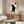 May include: Black cat poster with Japanese characters and red heart displayed above wooden sideboard in modern living room. Artwork shows silhouetted cat sitting upright on cream vintage-style background. Room features white curved chair, oval glass coffee table with colorful abstract design, and warm wood furniture against neutral walls.