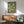 May include: Framed poster with green background featuring flock of white cranes in flight, displayed above wooden mid-century modern credenza with table lamp, next to sage green upholstered armchair with wooden frame, natural sunlight casting shadows across white wall in minimalist living room setting.