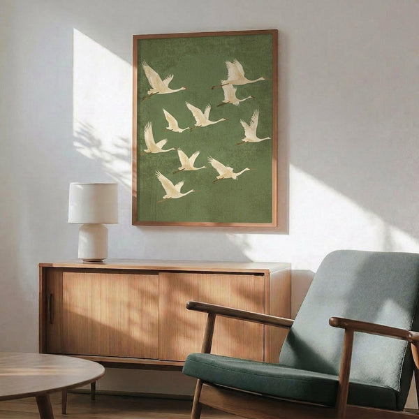 May include: Framed poster with green background featuring flock of white cranes in flight, displayed above wooden mid-century modern credenza with table lamp, next to sage green upholstered armchair with wooden frame, natural sunlight casting shadows across white wall in minimalist living room setting.