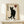 May include: Framed poster featuring black cat silhouette with yellow eyes on aged beige background, Japanese characters on left side, red heart with black character in upper right corner. Light wood frame sits on white shelf next to small white wrapped gift with evergreen sprig. Minimalist wall and decor styling.