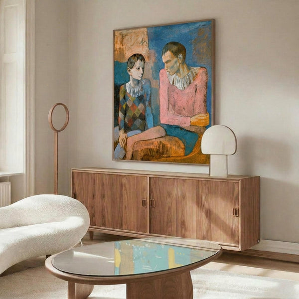 May include: Picasso's 1905 Two Acrobats artwork displayed above a walnut credenza in a modern living room. The painting shows two figures in harlequin costumes - one in diamond-patterned clothing, one in pink - against blue and coral backgrounds. White sculptural chair and glass-top coffee table complete the mid-century modern interior with warm wood tones and neutral walls.