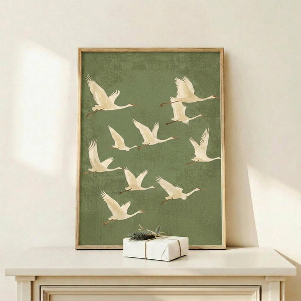 May include: Framed poster with green background showing nine white cranes in flight, displayed on light wood console table next to small wrapped gift with evergreen sprig. Natural wood frame, soft lighting creates gentle shadows on cream wall behind.