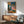May include: Framed reproduction of Picasso's 1905 painting showing two circus performers - a young boy in diamond-patterned harlequin costume and seated adult figure in pink garment with white ruff collar. Artwork displays muted blues, pinks, and earth tones in classical style. Print hangs above mid-century modern wooden credenza with ceramic table lamp, positioned next to sage green upholstered armchair with wooden frame in bright, naturally-lit room.